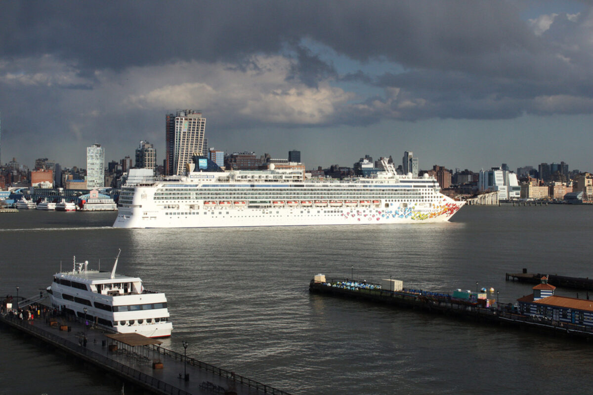 A white cruise ship passes on the Hudson under a dark, stormy sky, with Hoboken piers and a ferry in the foreground and the Manhattan skyline behind