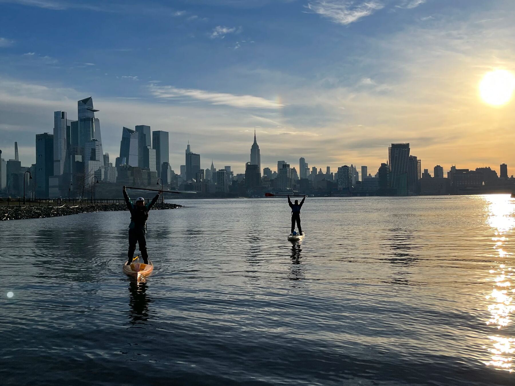 Two paddlers stand on boards with arms raised on calm water off Hoboken, the Midtown skyline behind them at sunrise